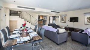 Een moderne woonkamer met een eethoek, grijze banken, een flatscreen televisie en een trap naar een keuken.