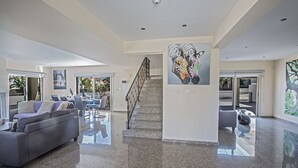 Een ruime woonkamer met een grijze bank, een eettafel en een trap naar een balkon.