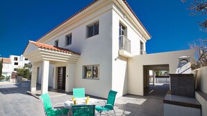 Een huis met twee verdiepingen, een balkon, een terras met tafel en stoelen, en een klein buitenkeuken.