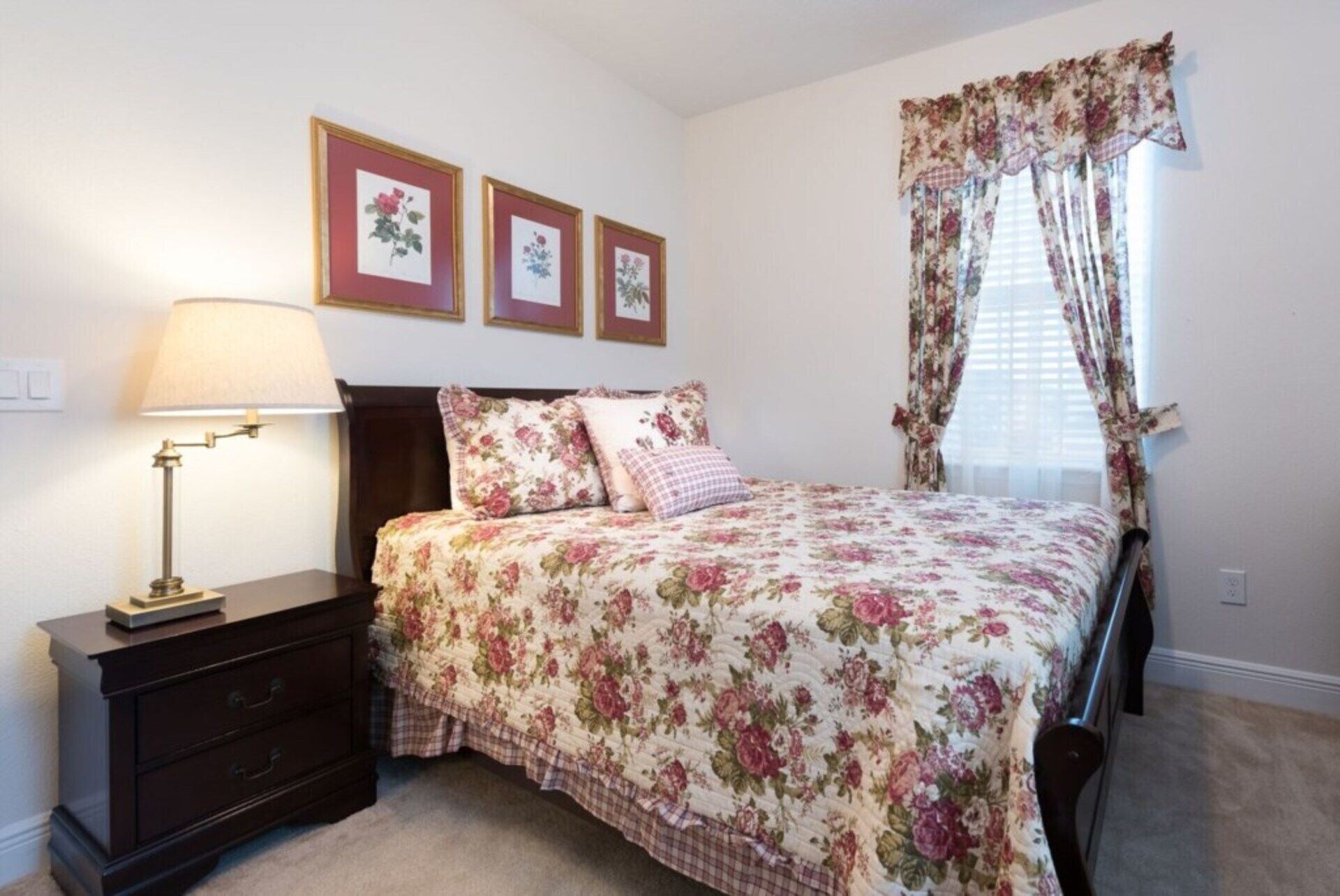 A bedroom with a bed, a nightstand, a lamp, a dresser, and floral curtains.