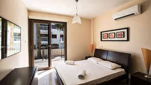 Habitación de hotel con cama, escritorio, silla, lámpara, aire acondicionado y vista al balcón exterior.