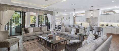 A spacious living area with a sofa set, coffee table, and chairs, a kitchen with white cabinets, and a ceiling fan.