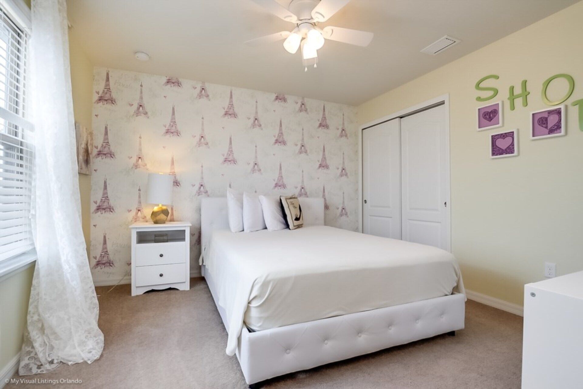 A bedroom with a bed, nightstand, and a ceiling fan. There is a door and a wall with the word "SHOP" and heart decorations.