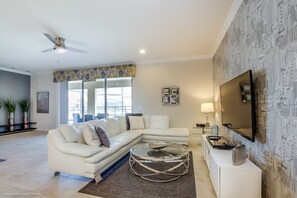 A modern living room with a sectional sofa, a glass coffee table, a flat-screen TV mounted on a textured wall, and a ceiling fan. - Villa in Windsor at Westside Resort 2799 (Kissimmee)