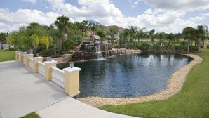 A landscaped pond with a waterfall, surrounded by palm trees and a paved walkway. - Townhome in Champions Gate Resort 2819 (Kissimmee)