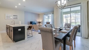 A modern dining area with a large table, comfortable chairs, a kitchen island, and a large window with curtains. - Townhome in Champions Gate Resort 2544 (Davenport)