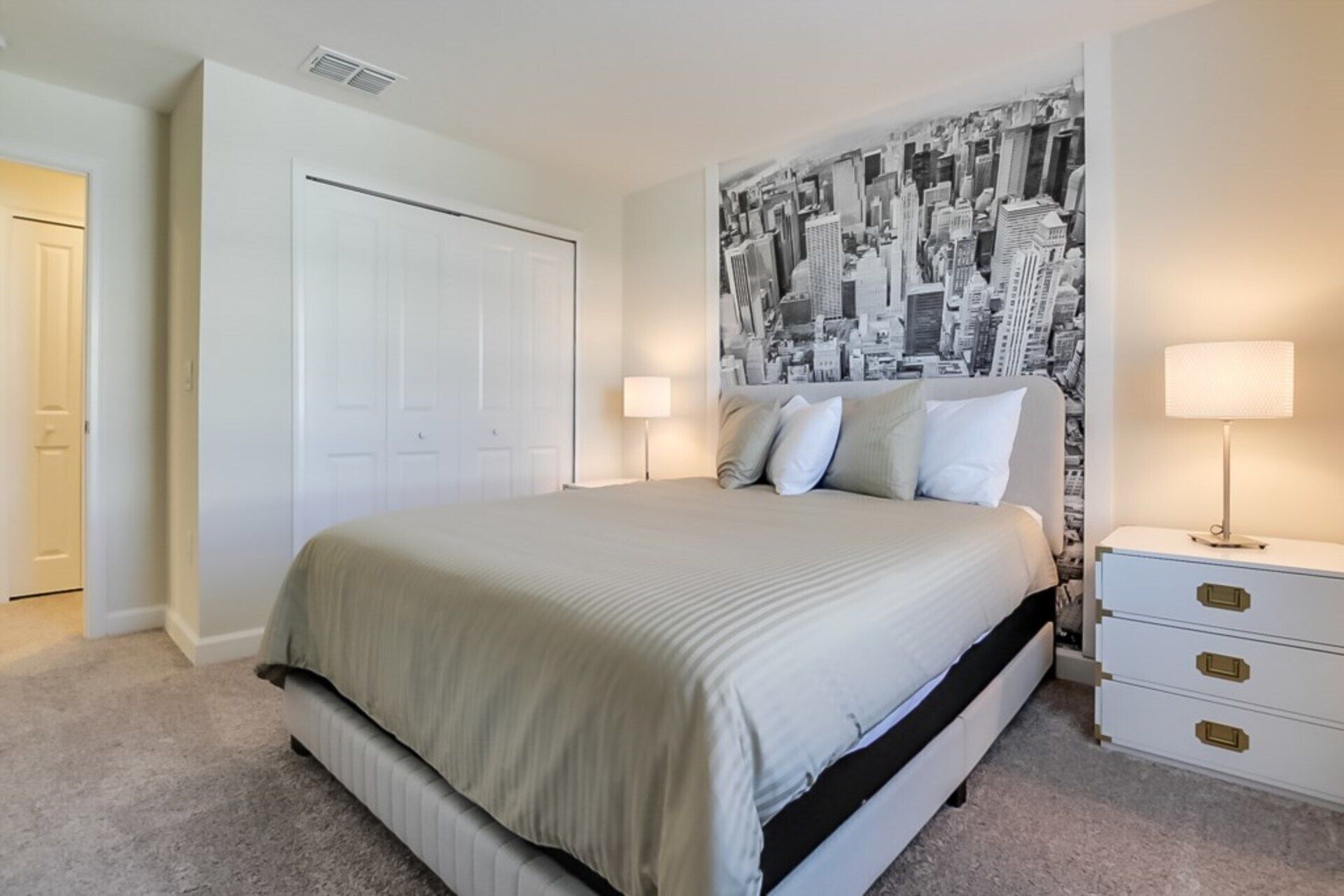A bedroom with a bed, a nightstand, a lamp, a wall with a cityscape mural, and a door.
