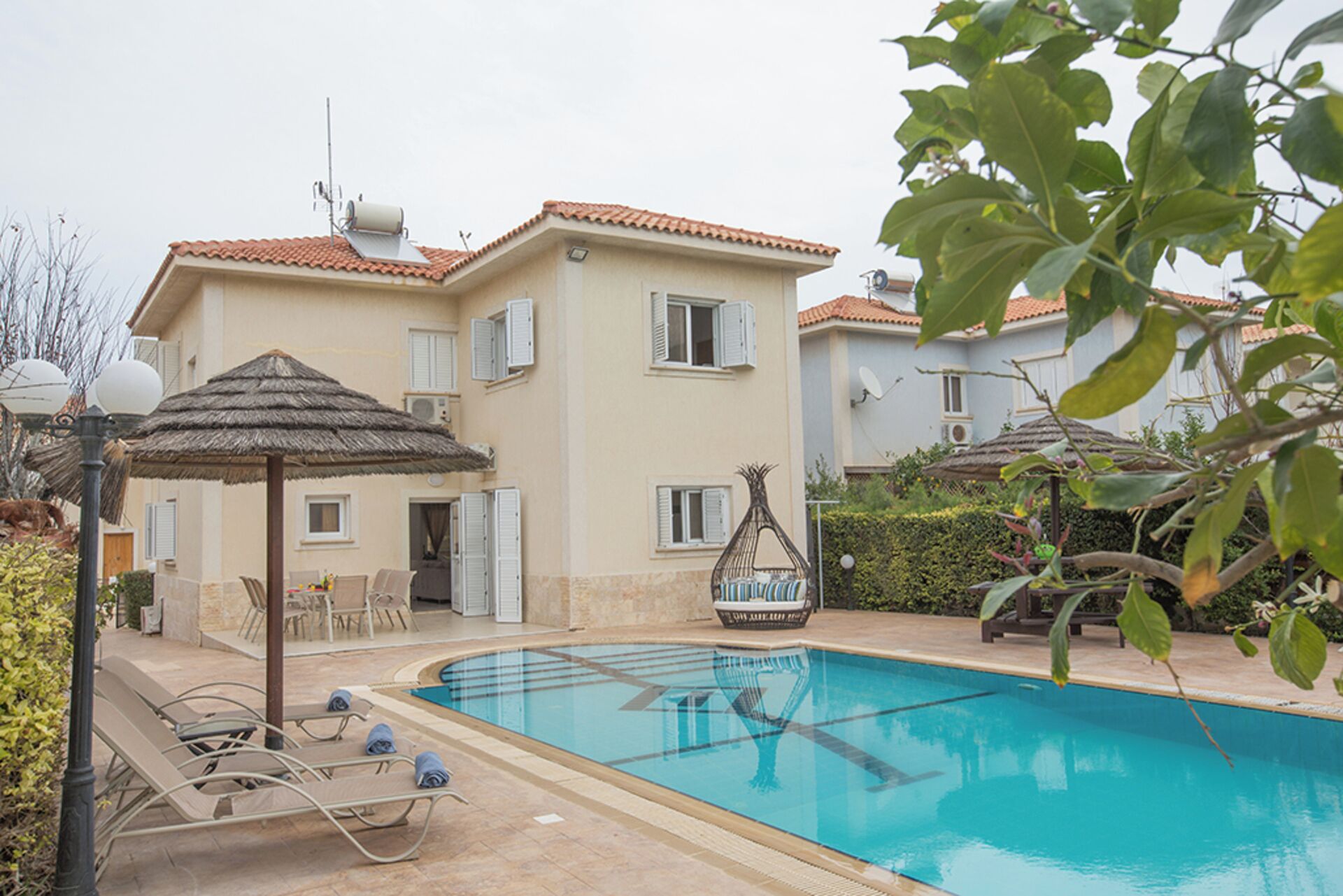 A swimming pool with lounge chairs, a thatched umbrella, and a building with a tiled roof.