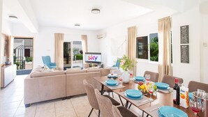 A modern living room with a sofa, dining table set for four, and a TV displaying the SKY channel. - Luxury Villa in Cyprus near Beach, Protaras Villa 1267 (Paralimni)