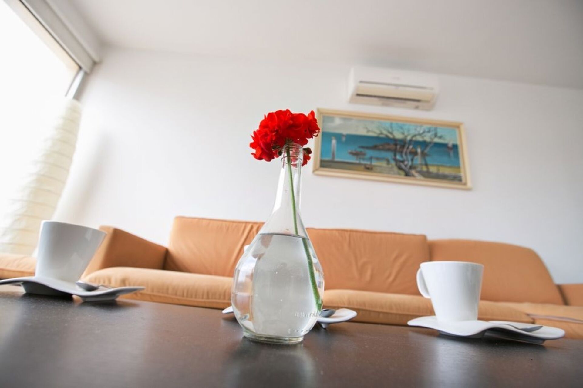 A room with a glass vase with red flowers on a table, a white cup, and a white saucer. There is a brown sofa and a painting on the wall.