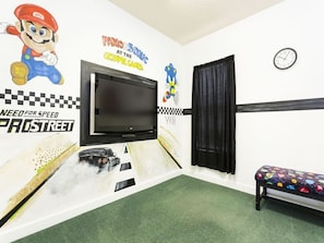 Ett rum med en stor TV, en Nintendo Wii-konsol, en vägg dekorerad med motiv från motorsport och en bänk med färgglada mönster.