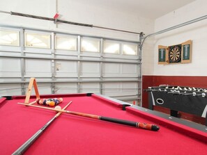 A pool table with a cue stick, billiard balls, and a foosball table in a room with a garage door and dartboards on the wall. - Villa in Windsor Palms Resort 1433 (Kissimmee)