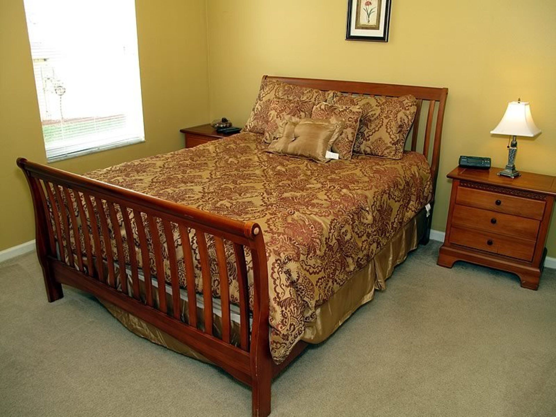 A bedroom with a wooden bed, a nightstand, a lamp, and a window with blinds.