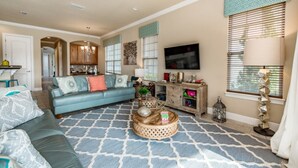 A living room with a blue sofa, a coffee table, and a TV. There is a kitchen in the background. - Luxury Villa With Majestic Golf Course Views, Reunion Resort, Villa Orlando 1795 (Kissimmee)