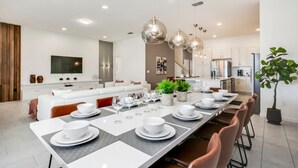 A modern kitchen with a long dining table set for six, white cabinets, stainless steel appliances, and pendant lighting. - Villa in Solterra Resort 2764 (Davenport)