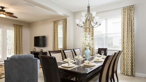 Comedor con una mesa de madera oscura dispuesta para seis personas, rodeada por seis sillas. Encima de la mesa hay un candelabro y un jarrón con flores, y una televisión de pantalla plana montada en la pared.