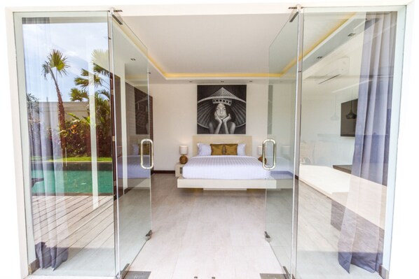 A modern bedroom with a glass door leading to a pool, a bed with white bedding, and a large framed artwork above the bed. - Private Villa in Seminyak 1156 (Seminyak)