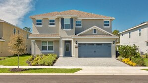 A two-story residential house with a front garage, a small porch, and a landscaped front yard with shrubs and a young tree. - Villa in Encore Resort at Reunion 2985 (Kissimmee)