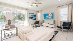 A spacious living room with a large white sofa, a glass coffee table, and a ceiling fan. There are two black chairs, a TV mounted on the wall, and a painting above the sofa. - Villa in Encore Resort at Reunion 2985 (Kissimmee)