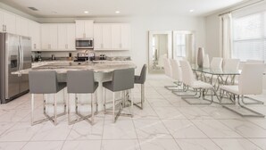 A modern kitchen with white cabinets, a stainless steel refrigerator, a microwave, and a gas stove. There is a marble countertop and a glass dining table with four chairs. - Villa in Encore Resort at Reunion 2985 (Kissimmee)