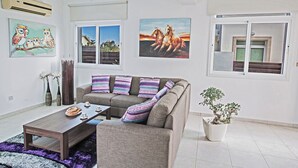 Un salón con sofá, una mesa de centro y una planta en maceta. Hay dos ventanas con cortinas, y un cuadro en la pared.