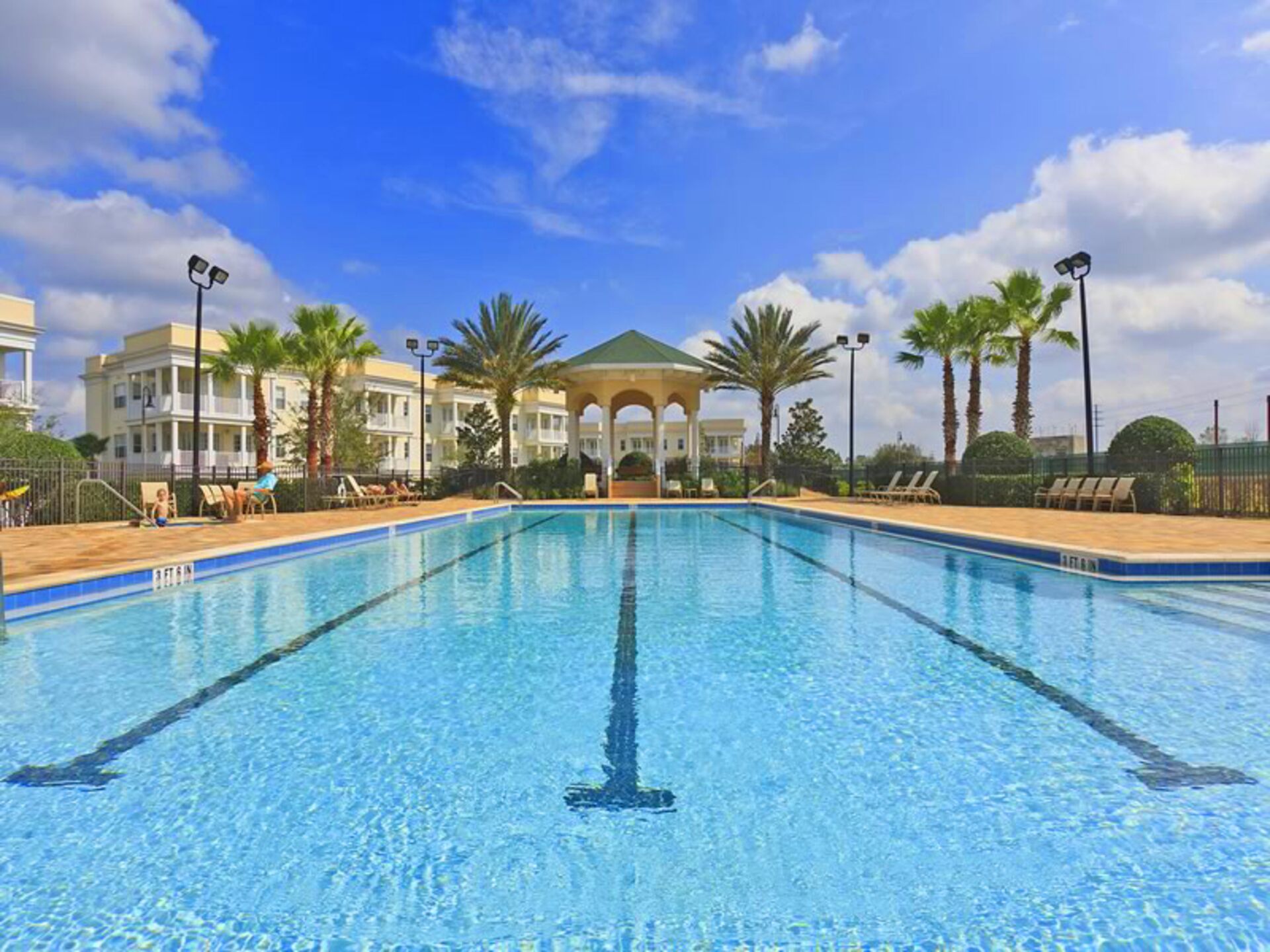A swimming pool with clear blue water, surrounded by lounge chairs and palm trees, with a building in the background.