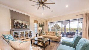 A spacious living room with a large sofa, a coffee table, and a television mounted on the wall. There is a ceiling fan, a large window with curtains, and a view of the outdoors.