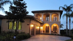A two-story house with a driveway leading to a gated entrance, surrounded by palm trees and a well-maintained lawn.