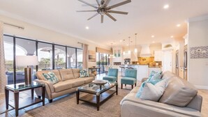 A spacious living room with a large window, a ceiling fan, and a kitchen area visible in the background.