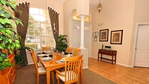 A dining area with a wooden table set for four, surrounded by chairs, a potted plant, and a view through a window with curtains. - Perfect 5 Bed Villa for Rent on Highlands Reserve Resort, Villa Orlando 1450 (Davenport)