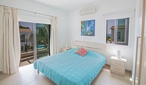 A bedroom with a bed, a nightstand, a painting, and a view of a pool and palm trees.