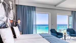 Quarto de hotel com uma cama grande, varanda com vista para o mar e área de estar com cadeiras e mesa.