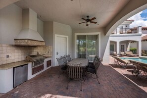 An outdoor patio with a grill, dining table, and chairs, a ceiling fan, and a view of a swimming pool and building in the background. - Villa in Reunion Resort & Spa 2976 (Kissimmee)