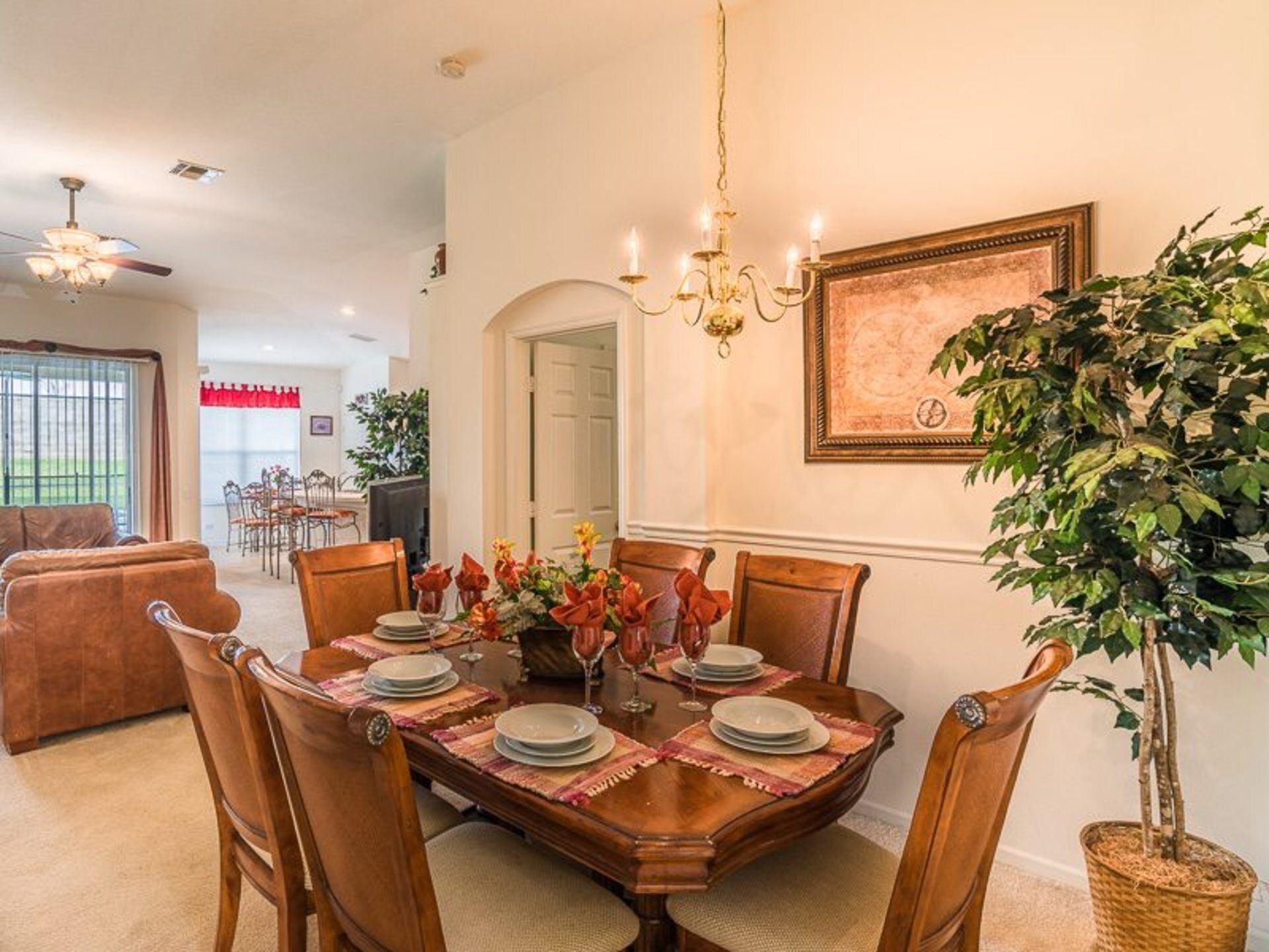 A dining room with a wooden table set for six, surrounded by chairs. A chandelier hangs above, and a large framed artwork is on the wall.