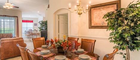 A dining room with a wooden table set for six, surrounded by chairs. A chandelier hangs above, and a large framed artwork is on the wall.