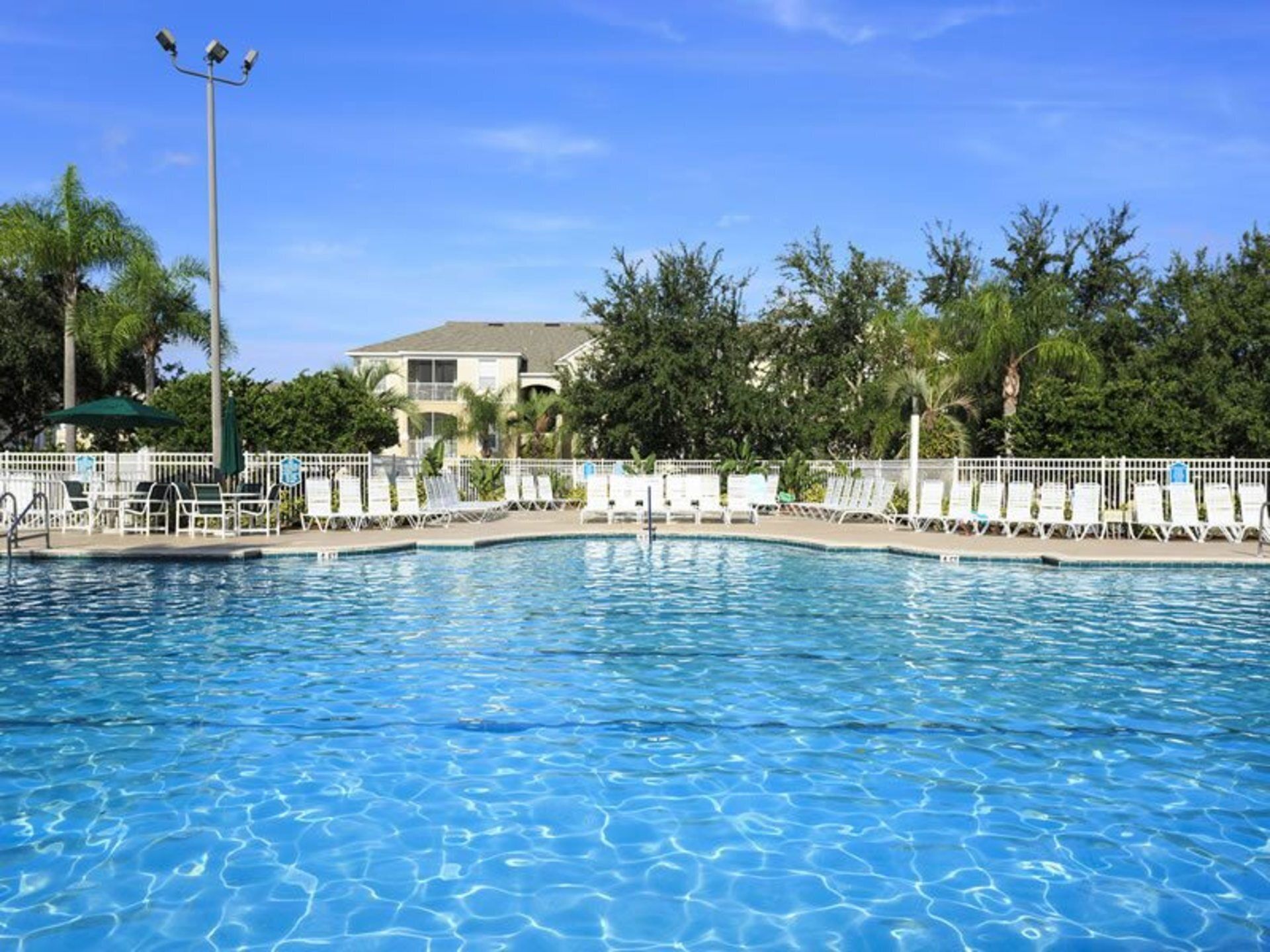 Piscina com espreguiçadeiras e cerca, cercada por árvores e um edifício ao fundo.