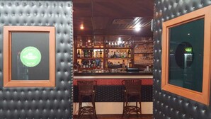 Una zona de bar con estantes llenos de botellas y vasos, dos sillas de mimbre y un certificado enmarcado en la pared.