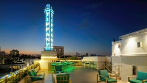 Eine Dachterrasse mit grünen Stühlen und Tischen, einer Neonreklame und einer Stadtansicht im Hintergrund.