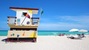 Ein Rettungsschwimmturm mit Hinweisschild für das Bad, Strandstühle und Sonnenschirme an einem Sandstrand.