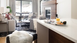 Habitación de hotel moderna con zona de comedor, sofá, mesa de centro y bandeja de desayuno con tostadas, aguacate y jugo de naranja.