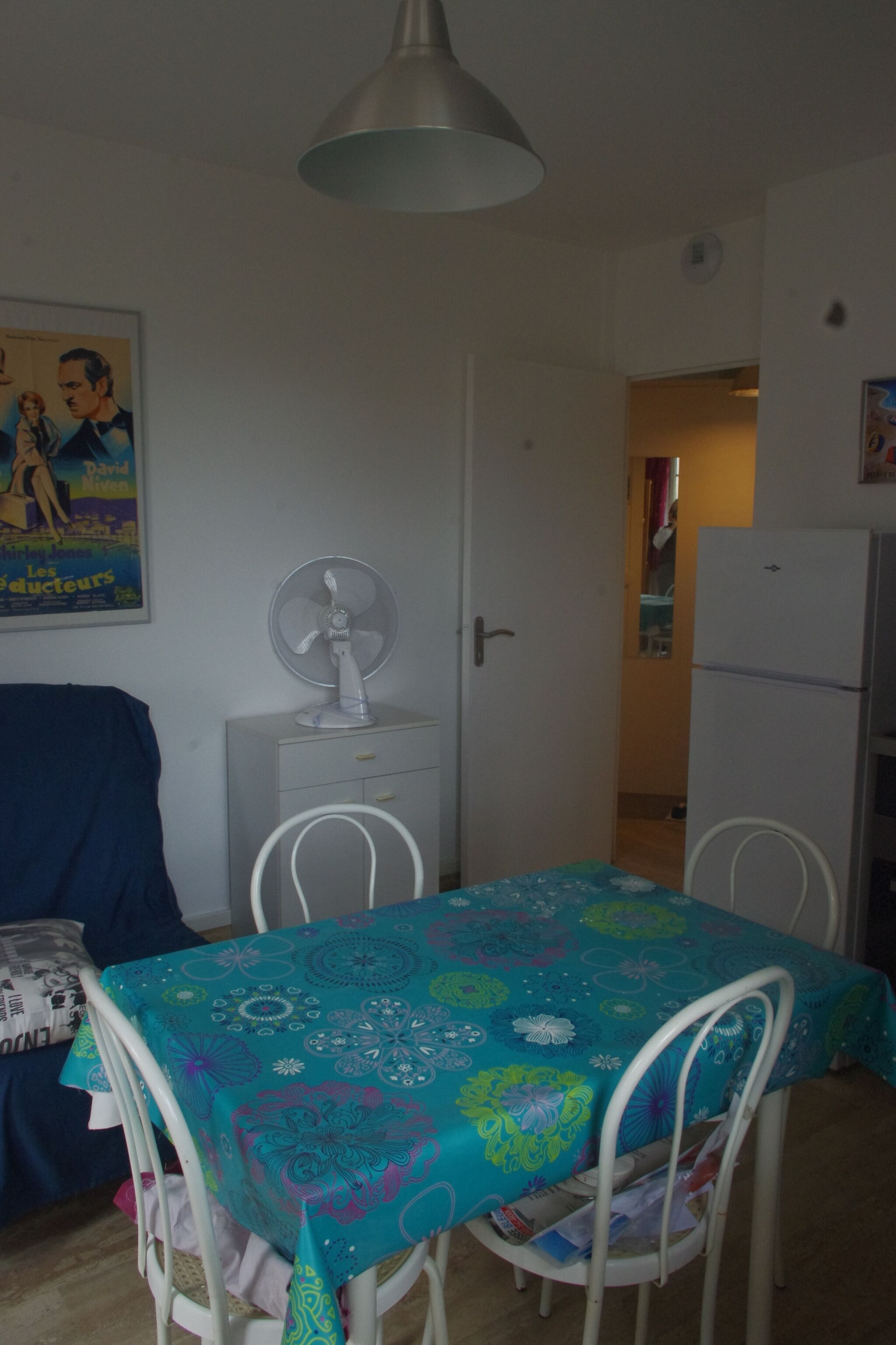 A room with a dining table covered with a colorful tablecloth, a white fan on a small table, a white refrigerator, and a door leading to another room.