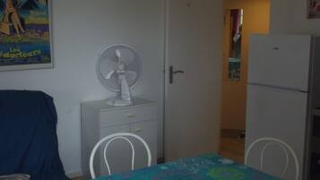 A room with a dining table covered with a colorful tablecloth, a white fan on a small table, a white refrigerator, and a door leading to another room.