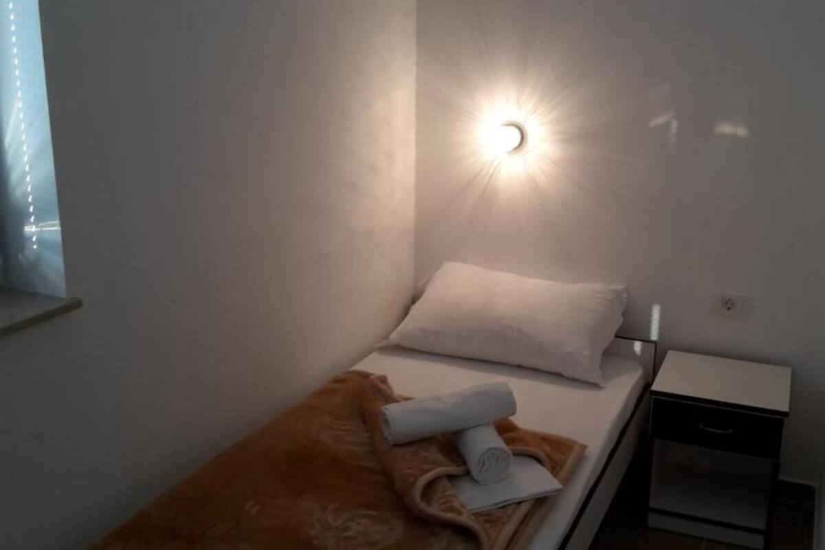 A single bed with a white pillow and a brown blanket, a small bedside table with a drawer, and a wall-mounted light fixture.