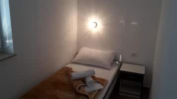 Un lit simple avec un oreiller blanc et une couverture marron, une petite table de chevet avec un tiroir et un luminaire fixé au mur.