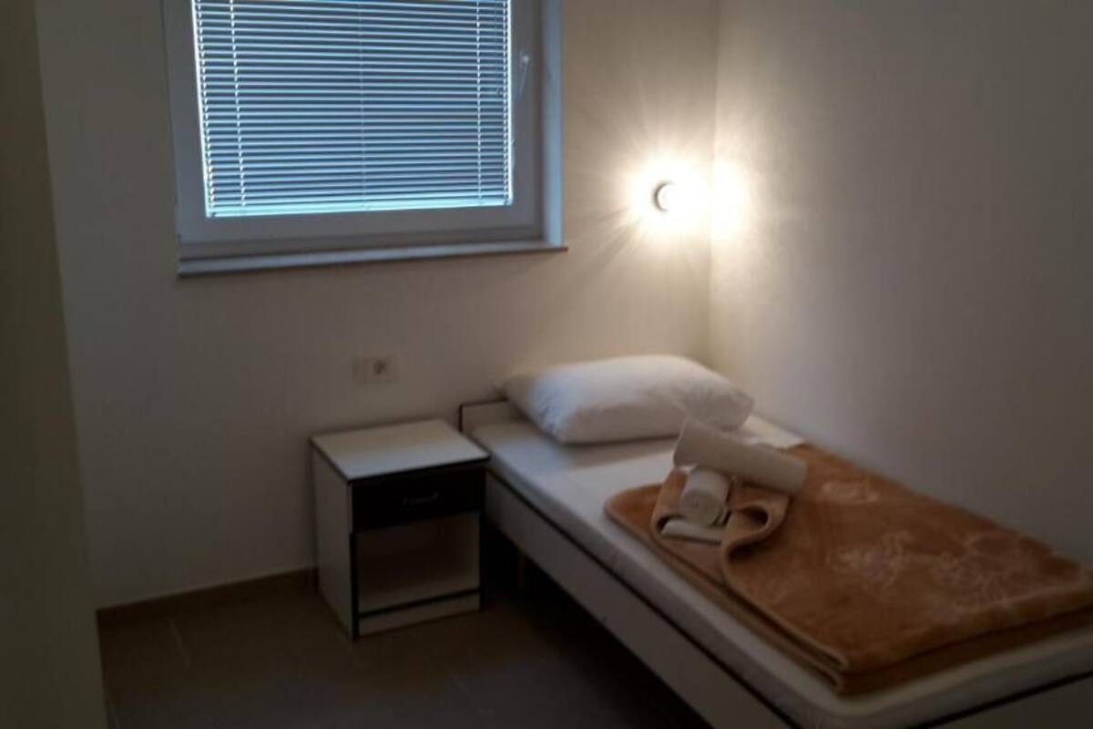 A small, single-bed room with a nightstand, a window with blinds, and a wall-mounted light.
