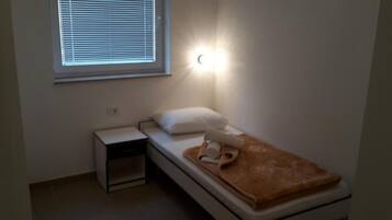 A small, single-bed room with a nightstand, a window with blinds, and a wall-mounted light.