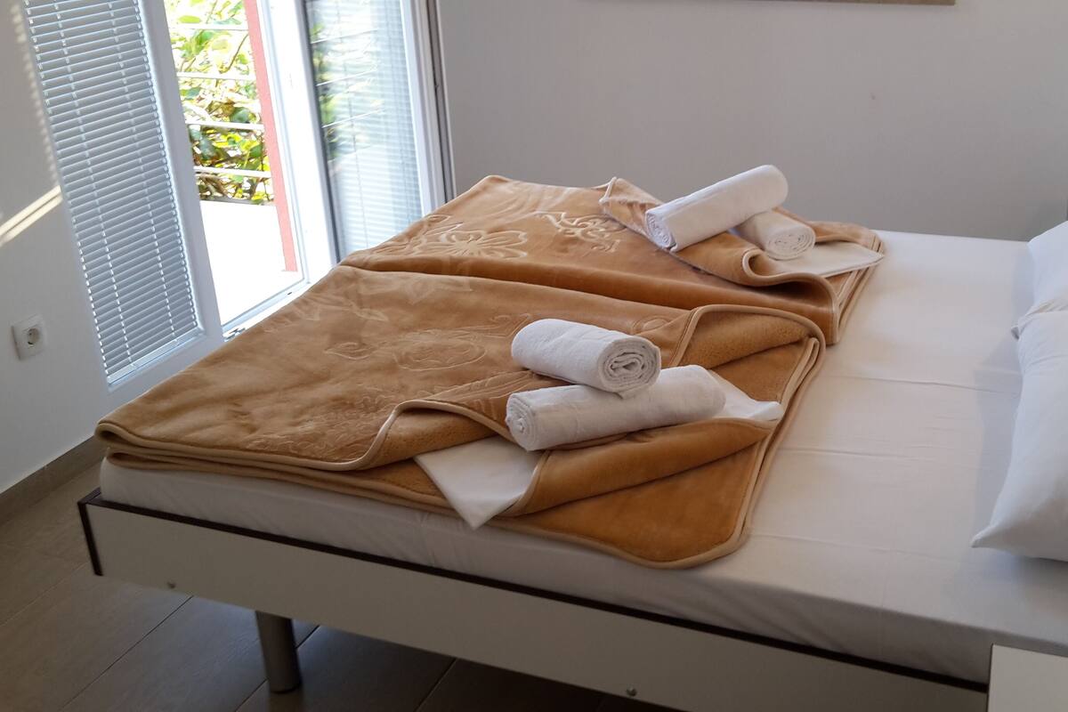 A neatly made bed with a brown blanket and two rolled white towels, a white pillow, and a window with blinds.