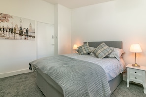 A bedroom with a bed, a nightstand, a lamp, a door, and a wall with a painting. - Hillcrest Lodge (Stevenage)