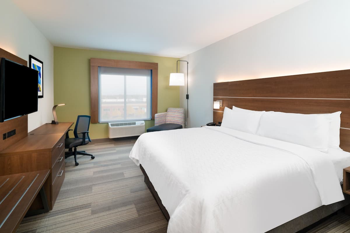 A hotel room with a large bed, a desk with a chair, a flat-screen TV, and a window with a view of the city.