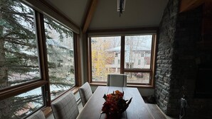 Un comedor con una mesa de madera, bancos y un florero. Amplias ventanas ofrecen vistas a árboles y edificios.